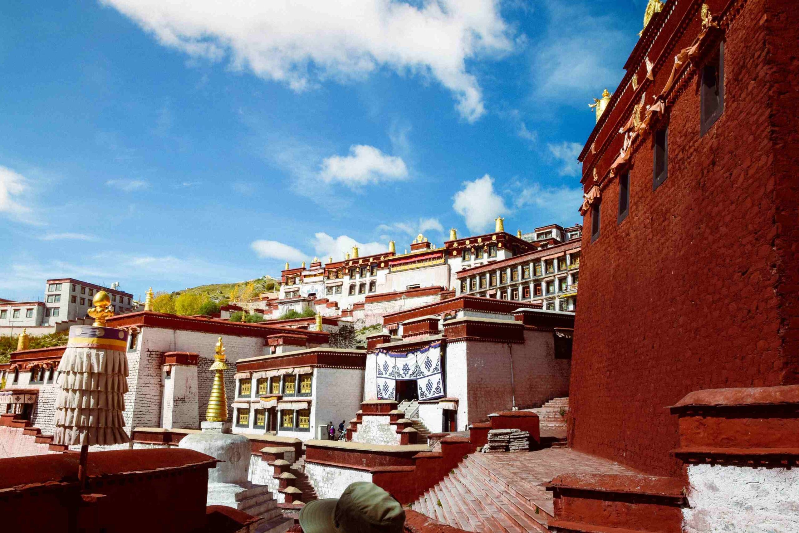 Private Tibet tour: The multi-tiered white and deep-red buildings of Ganden Monastery sprawling across a hillside under a blue sky in Lhasa, Tibet.